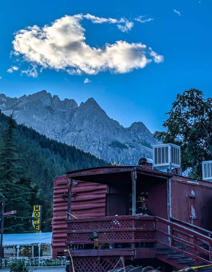 A rustic red railcar-style building with a small porch and exterior stairs sits beneath a bright blue sky, backed by dramatic rocky peaks and forested slopes. A fluffy white cloud floats above the mountains, and a yellow "Motel" sign is visible to the left, hinting at the location’s railroad-themed lodging.