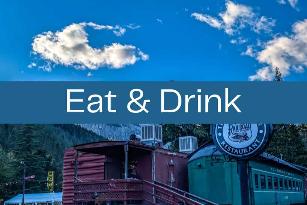 Vintage train cars repurposed as a restaurant, with a round “Railroad Park Resort Restaurant” sign and bright blue sky overhead. A bold blue banner across the center reads “Eat & Drink” in large white letters.