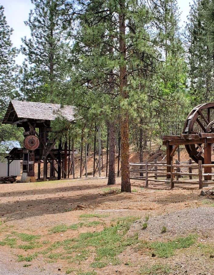 An old wooden mining structure and a large water wheel sit among tall pine trees in a forest clearing, part of a historic exhibit at Greenhorn Park. The rustic equipment is fenced off and surrounded by dirt paths and patches of grass, evoking a glimpse into Gold Rush-era industry.