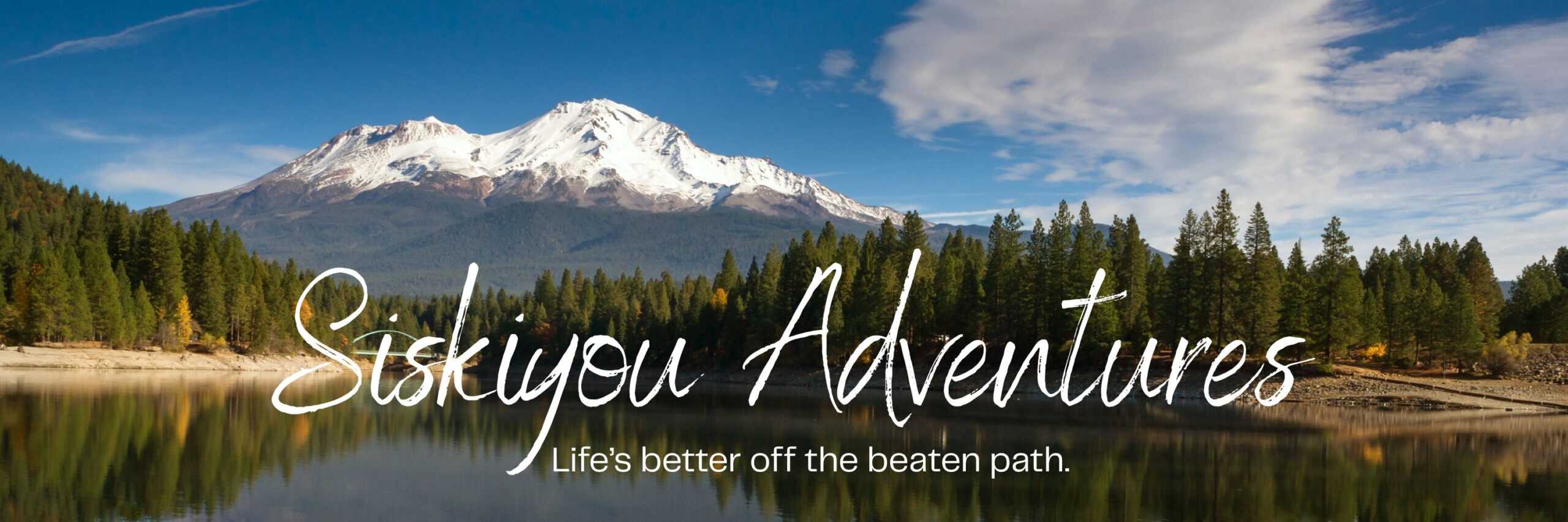 Scenic view of a snow-capped Mount Shasta reflected in a calm lake, framed by dense evergreen forest under a bright blue sky. Overlaid text reads “Siskiyou Adventures” with the tagline “Life’s better off the beaten path,” evoking outdoor exploration and nature travel.