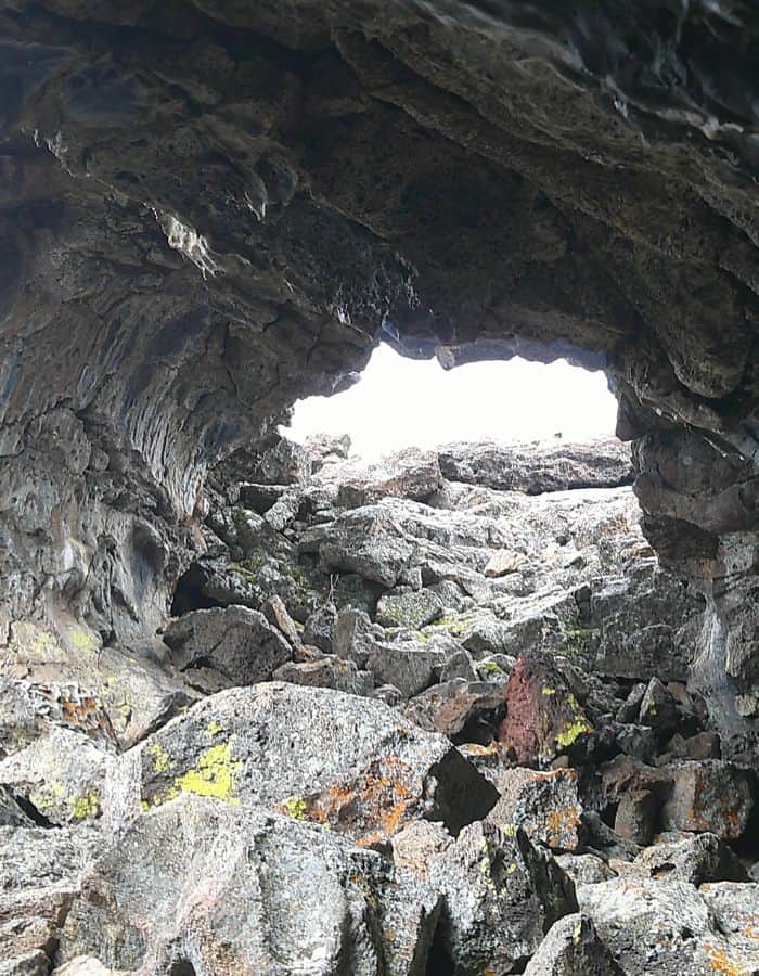 Looking out from the inside of a rugged lava tube, jagged volcanic rocks form the floor and walls, with a natural opening at the top revealing a bright, overcast sky. Bits of green and orange lichen cling to the dark, weathered stone, highlighting the cave's raw, natural texture.