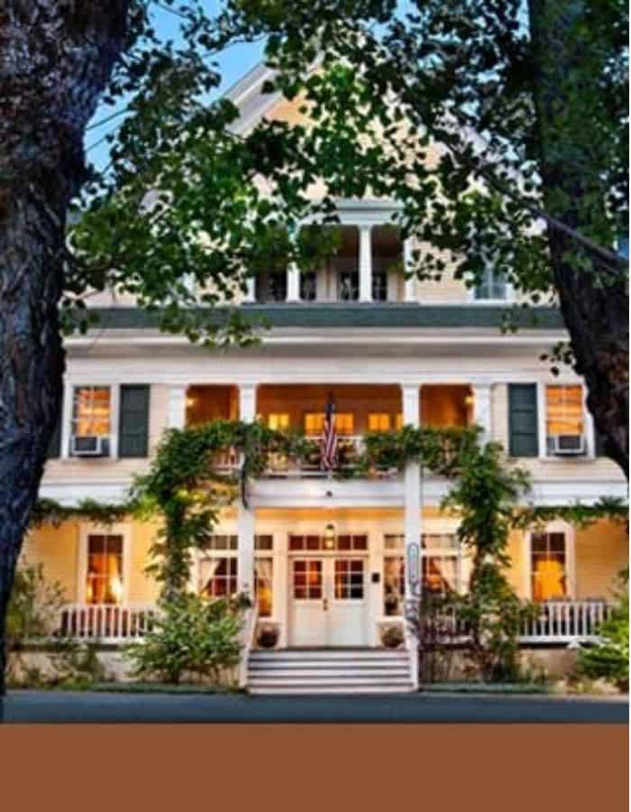 A charming three-story inn with white siding and green shutters is warmly lit from within at dusk, framed by two large trees. The hotel features a welcoming front porch draped in greenery and an American flag above the entrance, giving it a cozy, historic feel.