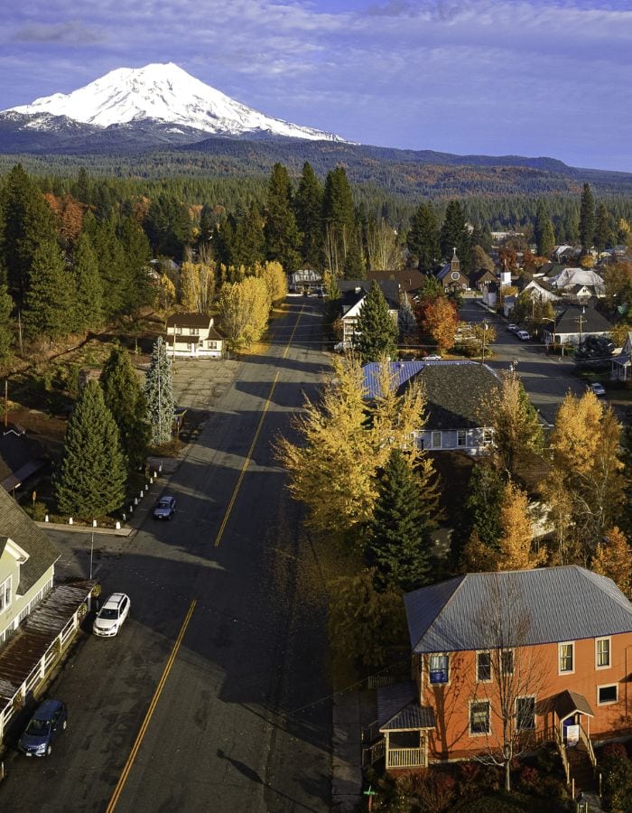 A quiet small-town street in McCloud, California, is lined with autumn-colored trees and quaint houses, leading toward a distant view of Mount Shasta capped in snow. The early morning or late afternoon sunlight casts a golden hue over the town, with long shadows stretching across the road.