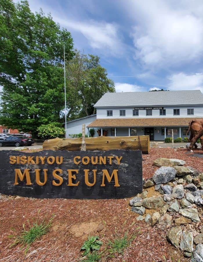 The Siskiyou County Museum building sits behind a large black and gold sign that reads “Siskiyou County Museum,” with a log and stone landscaping feature in front. The museum is housed in a light gray, two-story structure with a brown shingled awning and surrounded by lush greenery under a partly cloudy sky.
