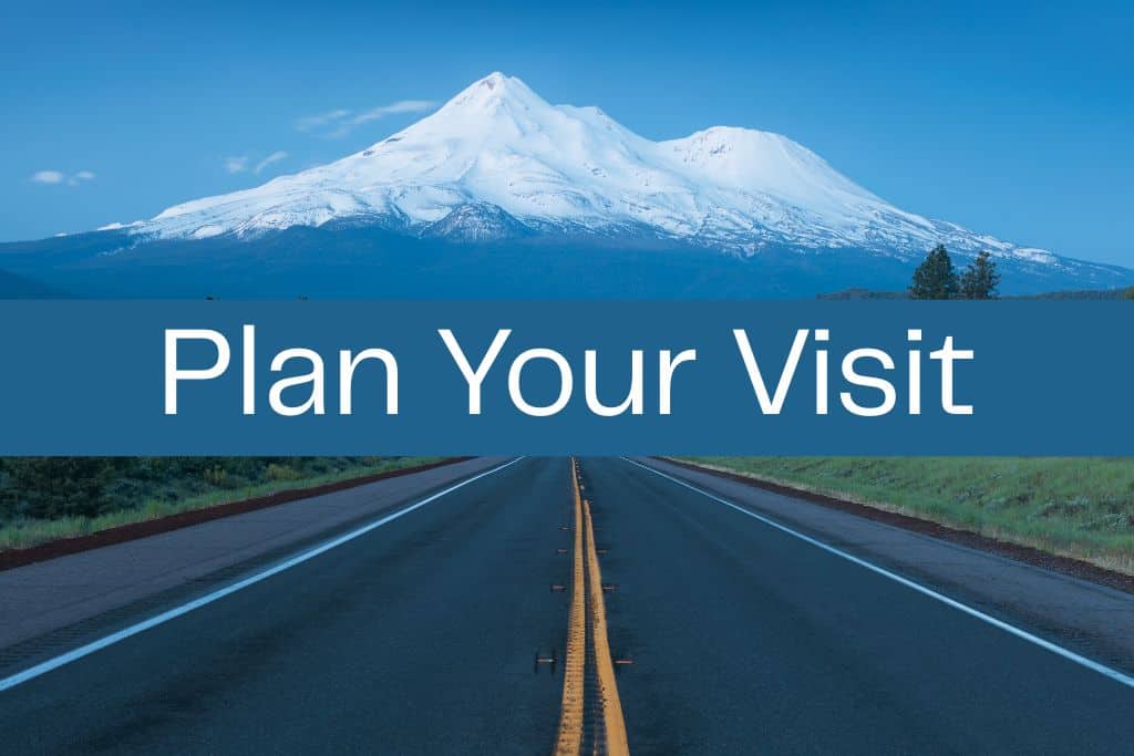 Open highway leading toward a snow-covered Mount Shasta under a clear blue sky. A wide blue banner across the center reads “Plan Your Visit” in large white letters.