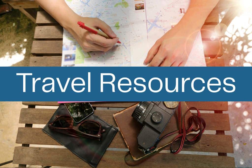 Person marking a location on a city map at an outdoor wooden table, with sunglasses, a camera, and a notebook laid out beside them. A bold blue banner across the image reads “Travel Resources” in large white text.