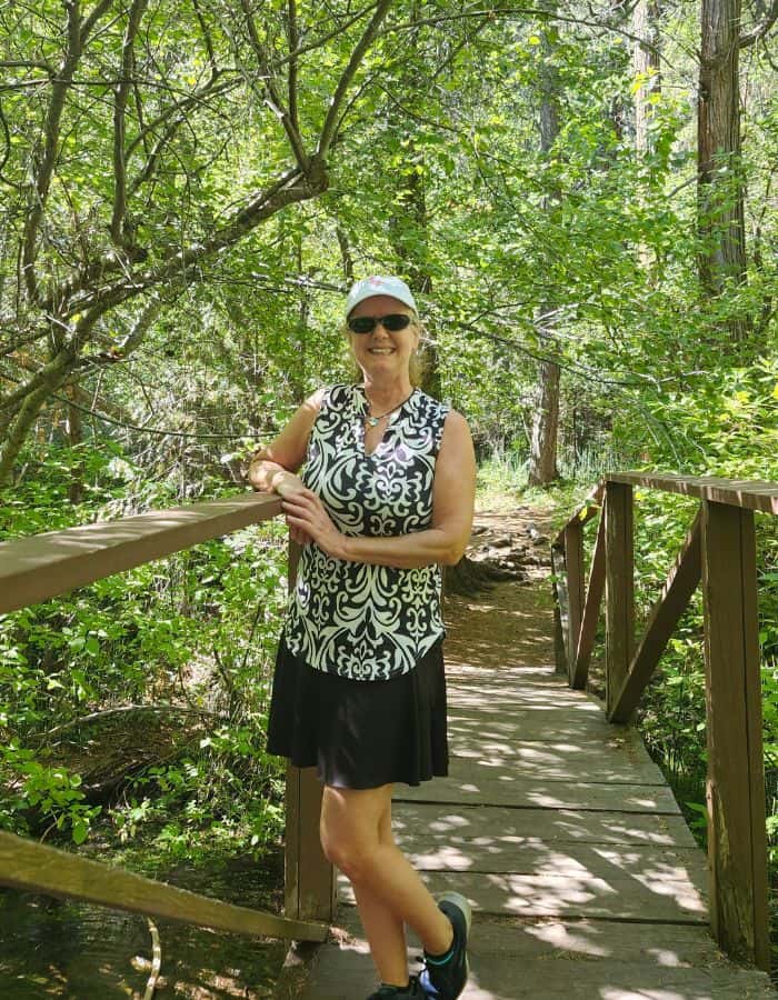 A woman wearing sunglasses, a patterned sleeveless top, black skirt, and white cap smiles while standing on a small wooden footbridge in a lush forest. Sunlight filters through the green canopy, casting dappled light on the path and stream below.