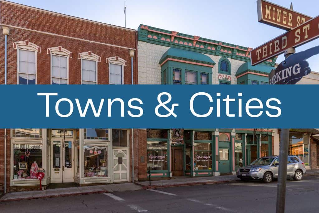 Quaint downtown street with historic brick and painted buildings, including shops and a restaurant. A blue banner across the center reads "Towns & Cities" in large white letters, with old-fashioned street signs for Miner Street and Third Street visible in the corner.
