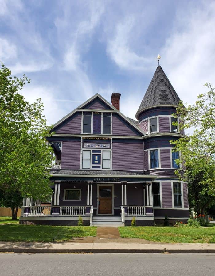 A grand Victorian house painted deep purple with white trim and gold accents stands on a well-kept lawn. The home features a prominent round turret with a pointed roof, a large front porch with ornate railings, and the name “Gillis Est. 1895” displayed above the entrance.