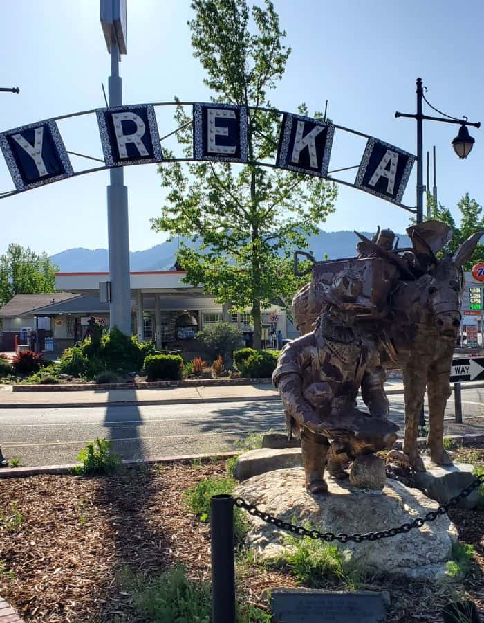 A metal archway spelling out “YREKA” stands over a roadside sculpture of a gold miner crouched beside a pack mule, set against a backdrop of gas stations, businesses, and distant mountains. The bronze statue captures Yreka’s Gold Rush heritage in a small landscaped plaza under clear blue skies.