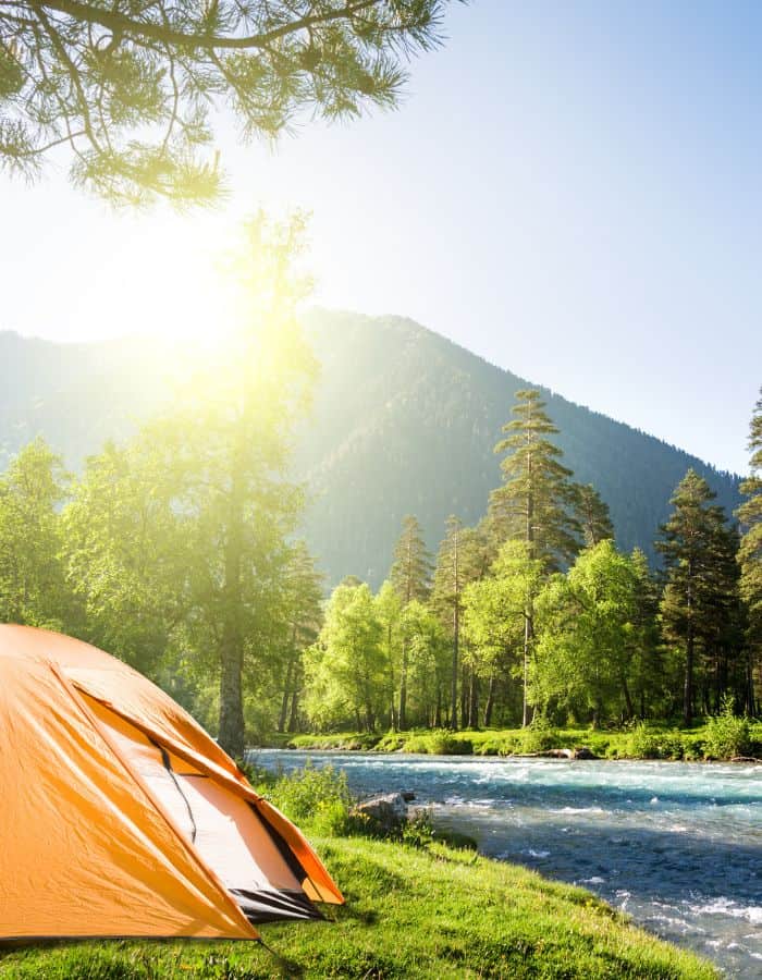 A bright orange tent is pitched beside a sparkling river surrounded by lush green trees, with sunlight streaming through the forest and illuminating a nearby mountain. The peaceful outdoor scene captures a perfect day for camping in nature.