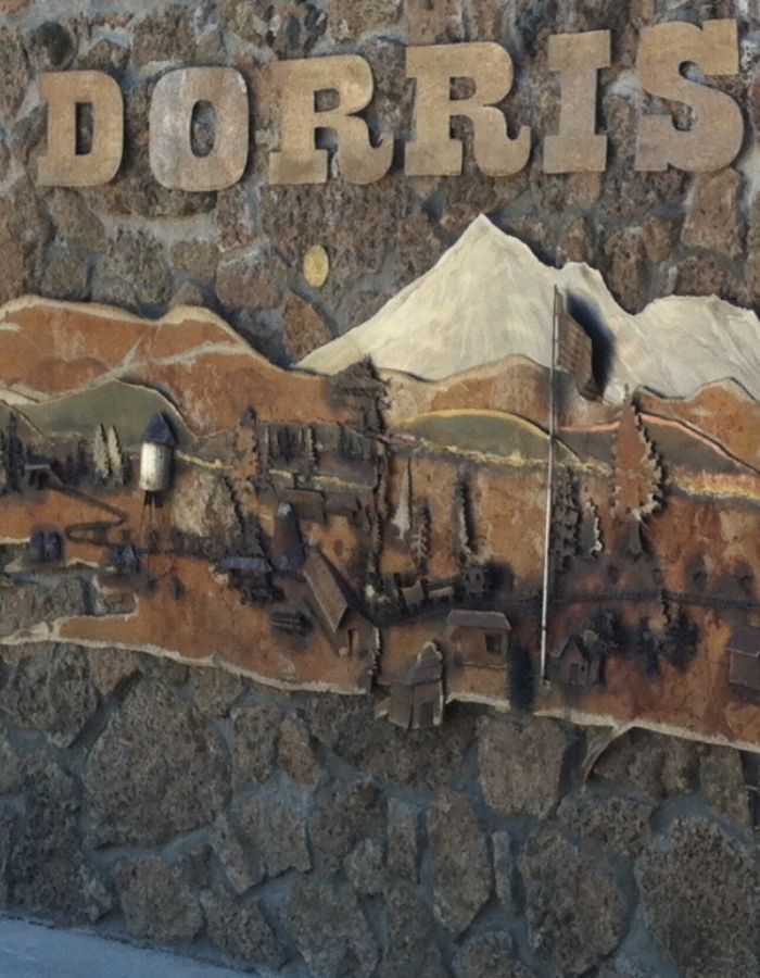 Rustic stone mural in Dorris, California, featuring a raised metal and wood relief of a small rural town with trees, houses, a water tower, and an American flag, set against the backdrop of a large snow-capped mountain. The word “DORRIS” is displayed in bold wooden letters above the scene.