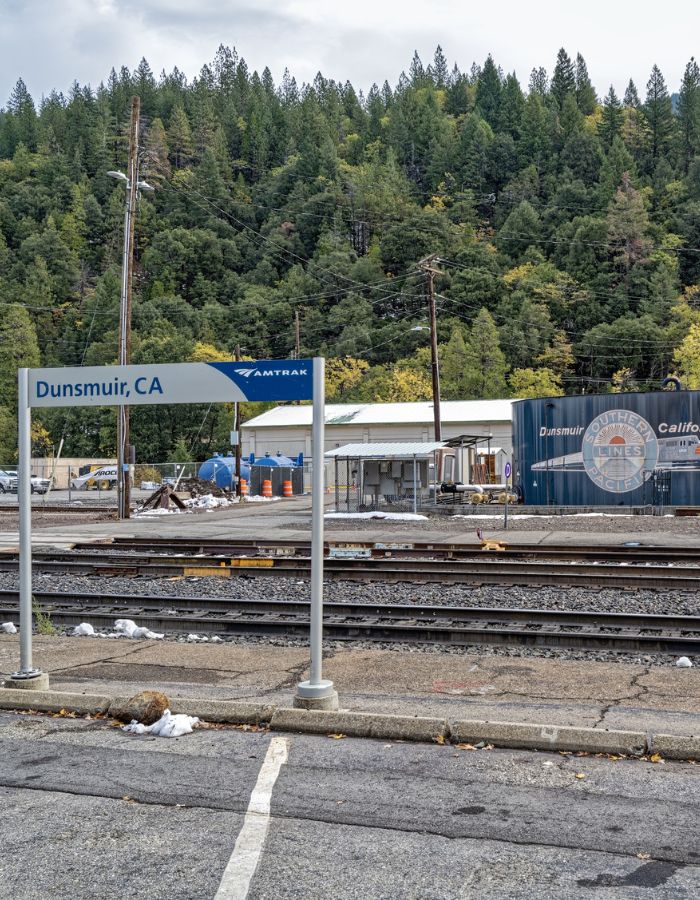 Amtrak station sign for Dunsmuir, CA in the foreground, with train tracks and an industrial area behind it, including construction barrels, storage tanks, and a large container marked “Southern Pacific Lines” and “Dunsmuir, California.” A dense forest of tall evergreen trees covers the hillside in the background.