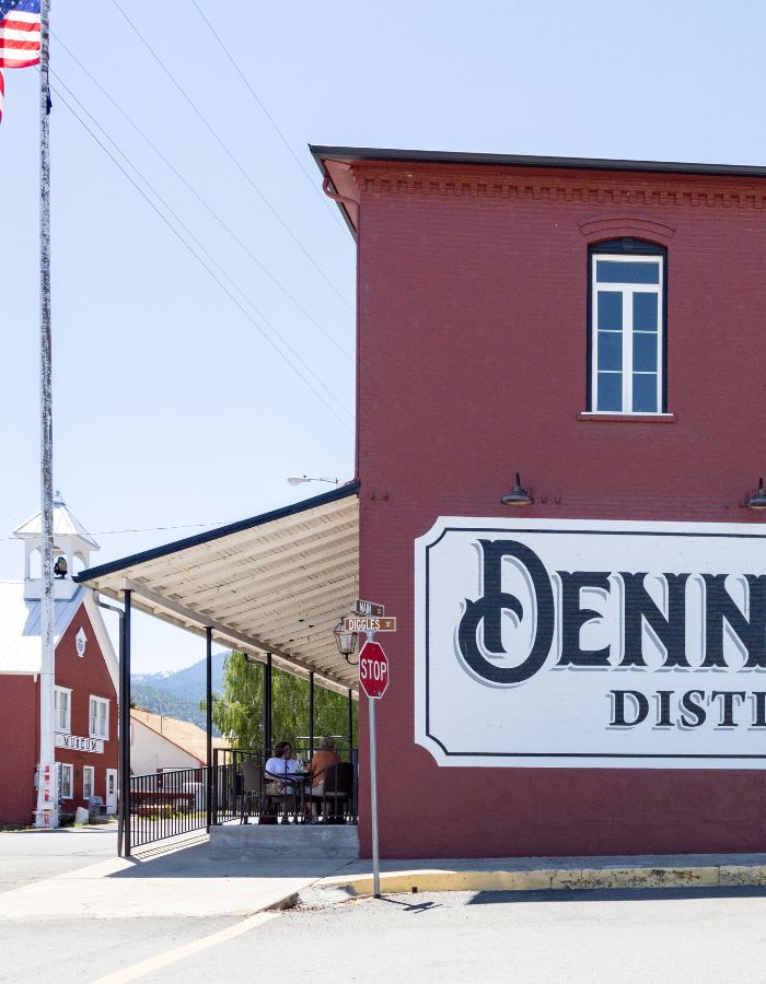 Corner view of a red brick building with a large painted sign reading “DENN” on the side, part of a larger sign for Denn Distillery. A covered patio with people sitting at tables extends from the building, and a street sign for “Diggles St” and a stop sign are at the corner. In the background, another red building labeled “MUSEUM” features a white bell tower and American flag, with tree-covered hills and blue sky beyond.