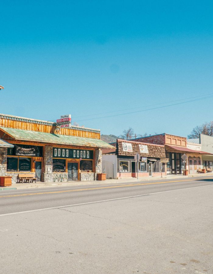 Sunny downtown street in Fort Jones, California, featuring a row of rustic storefronts including the prominent “Five Marys” restaurant and steakhouse with stone and wood paneling. Signs advertise cocktails, beer, and a live performance, while adjacent buildings have vintage-style signage and shingled awnings, all set against a clear blue sky and distant hills.