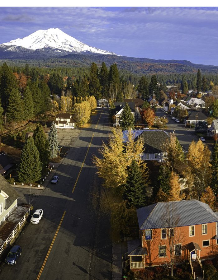 A quiet small-town street in McCloud, California, is lined with autumn-colored trees and quaint houses, leading toward a distant view of Mount Shasta capped in snow. The early morning or late afternoon sunlight casts a golden hue over the town, with long shadows stretching across the road.