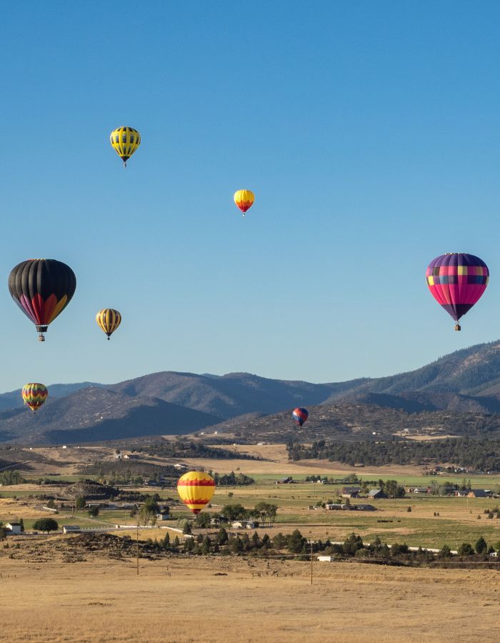 A group of colorful hot air balloons floats above a rural landscape in Montague, California, with open fields, scattered homes, and rolling hills in the background. The clear blue sky and low sunlight create a vivid, peaceful atmosphere as the balloons drift over the countryside.