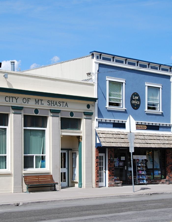 Street view of downtown Mount Shasta featuring two adjacent buildings—one cream-colored with green accents labeled “City of Mt. Shasta,” and another painted blue housing a “Law Office” and a shop called “Stage Quest.” A bench sits outside the city building, and a sandwich board sign with posters is displayed on the sidewalk.