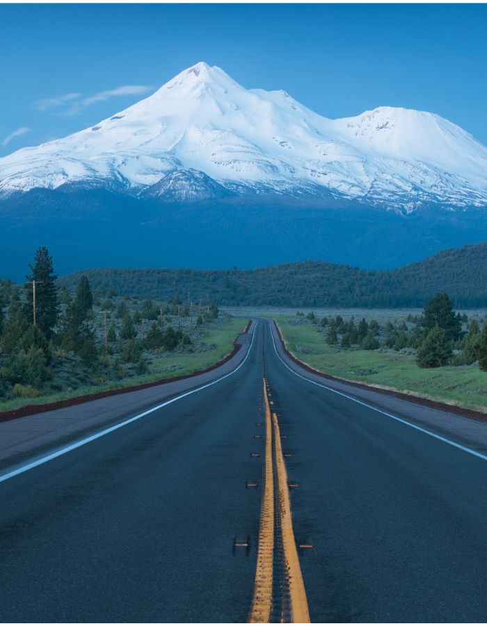 Long, straight highway leading through a forested valley toward the towering, snow-covered peak of Mount Shasta under a soft blue sky. The road’s yellow centerline guides the eye directly to the mountain, creating a dramatic and scenic composition.