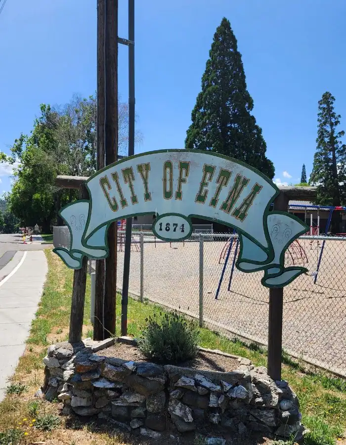 A rustic welcome sign reading "City of Etna 1874" stands on a stone base beside a sidewalk, with a school playground and tall evergreens visible in the background under a clear blue sky.